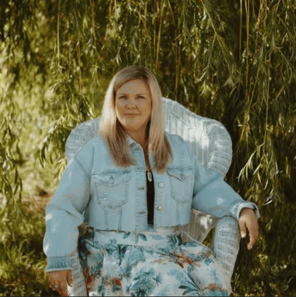 A woman with blonde hair in a denim jacket and floral skirt relaxes on a white wicker chair outdoors, surrounded by lush foliage, savoring the peaceful atmosphere after her Soulful Mentorship session.